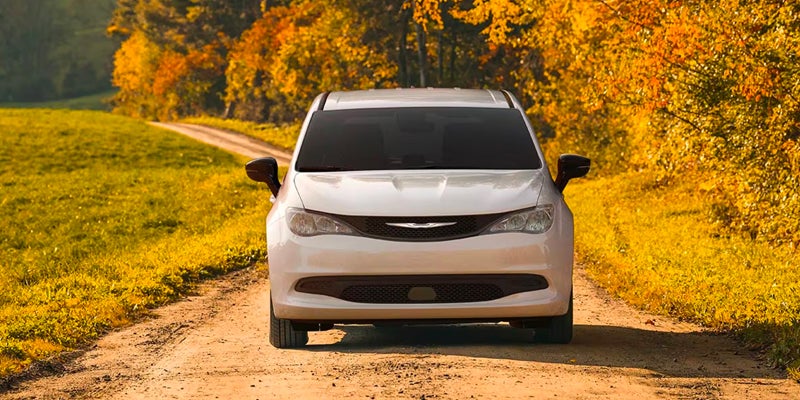 Front view of a white minivan parked on a rural dirt road surrounded by trees with autumn-colored foliage