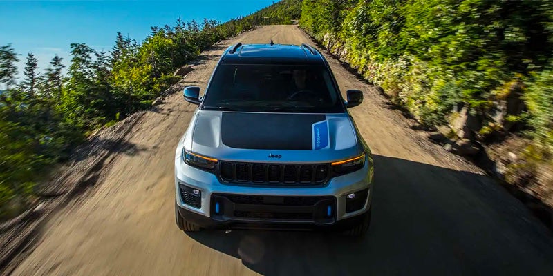 Silver Jeep Grand Cherokee with a black hood and blue stripe driving up a dirt road surrounded by green trees