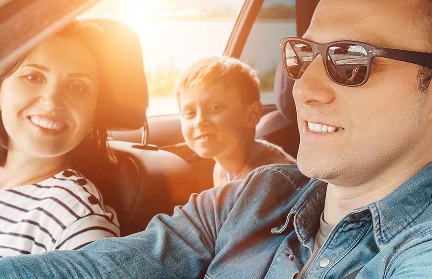 Smiling family in a car, bathed in bright sunlight; dad driving, mom next to him, and a child in the back seat