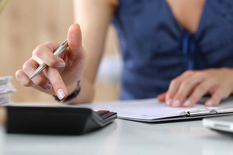 Close-up of a person holding a pen while reviewing documents and using a calculator on a desk