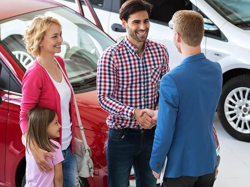 Smiling family at a car dealership, with the dad shaking hands with a salesman