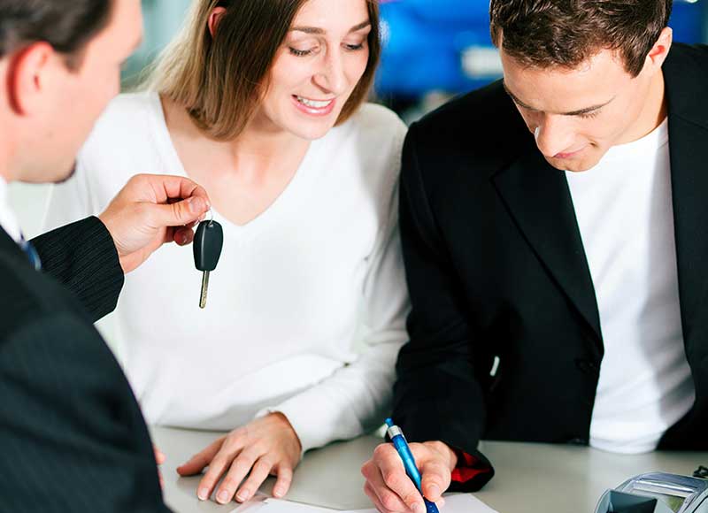 A man hands car keys to a happy woman as another man signs paperwork