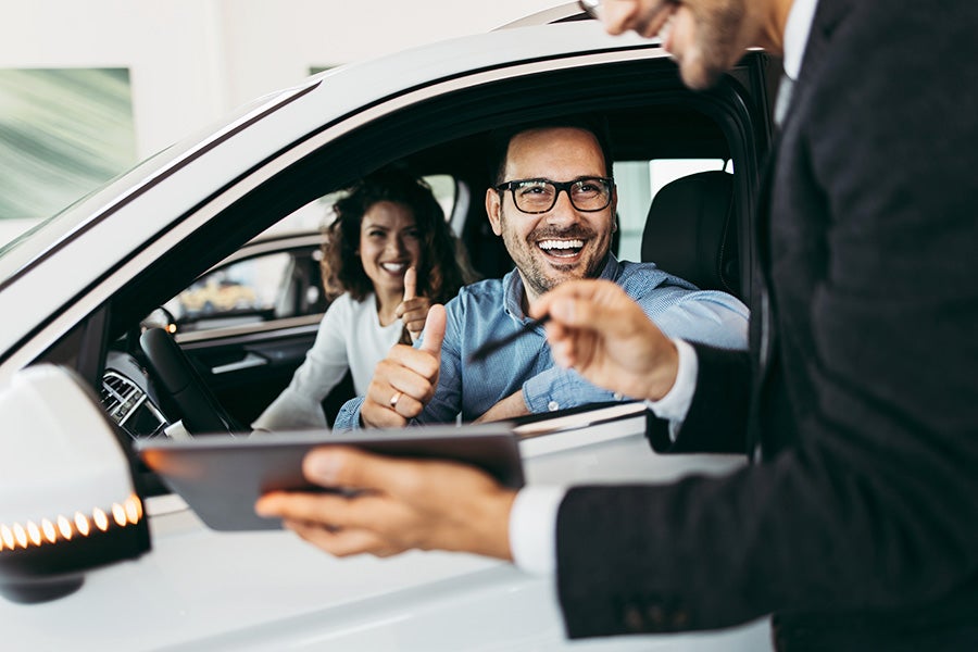 Happy couple in a new car giving thumbs up to a salesman holding a tablet