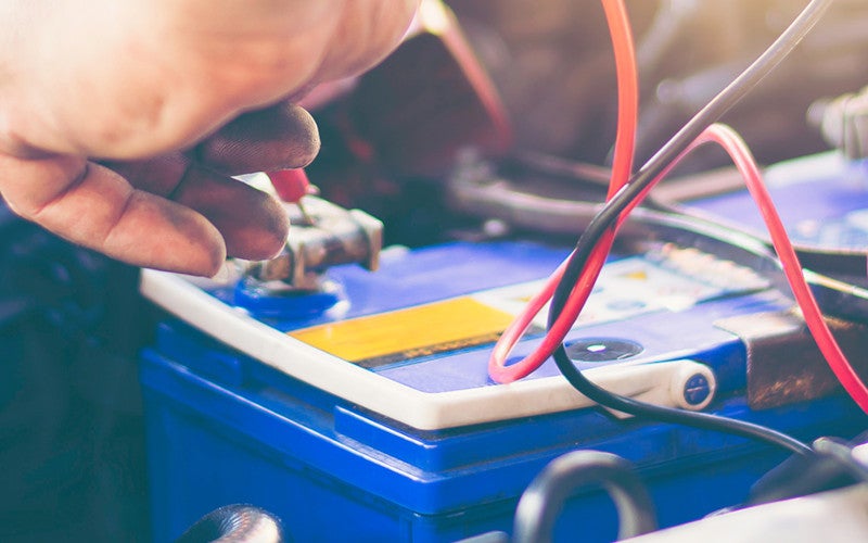 Close-up of a vehicle battery during maintenance, with a hand connecting testing cables to the battery terminals inside an engine compartment
