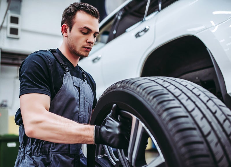 Automotive technician installing or inspecting a tire and wheel on a white SUV inside a vehicle service and maintenance facility
