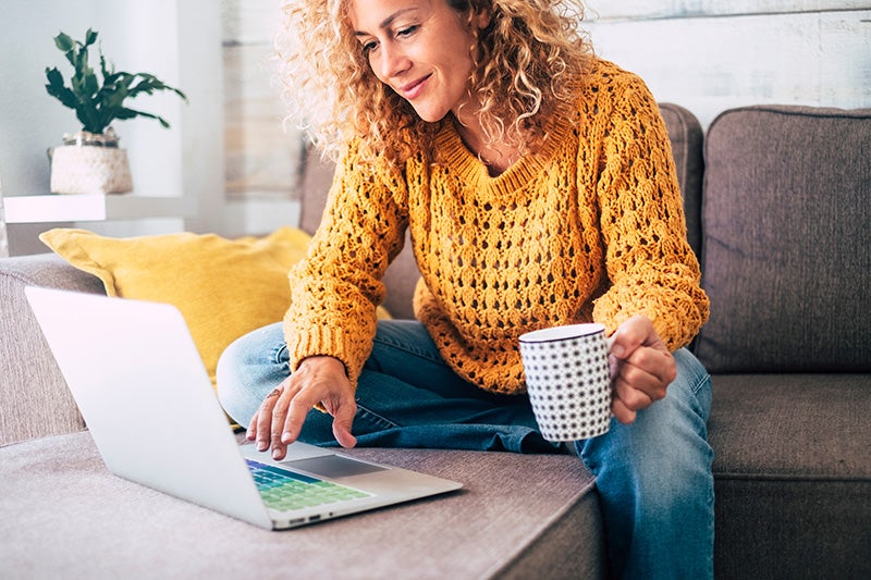 Curly-haired woman in a yellow sweater sits on a couch, working on a laptop and holding a mug
