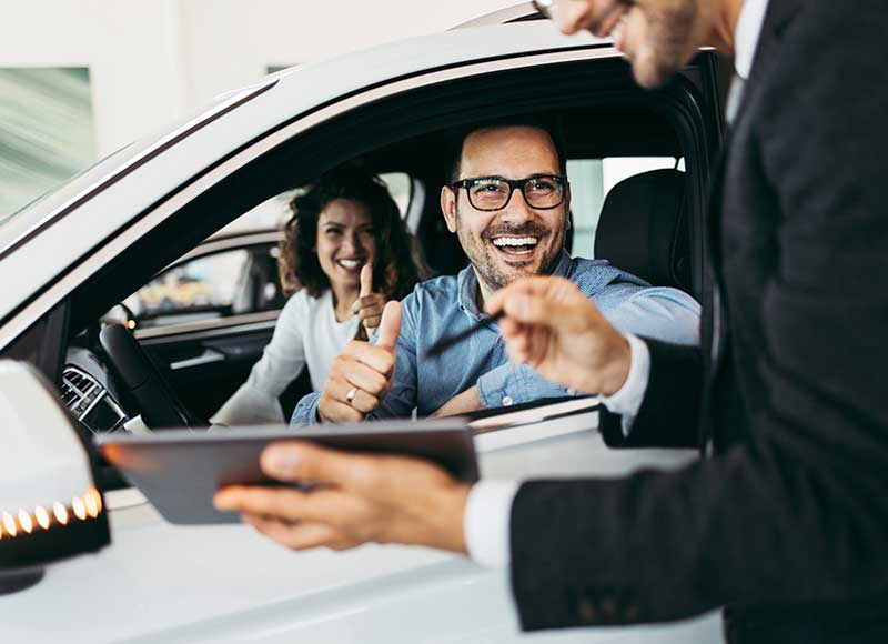 Happy couple in a new white car, smiling and giving thumbs-up to a salesperson holding a tablet